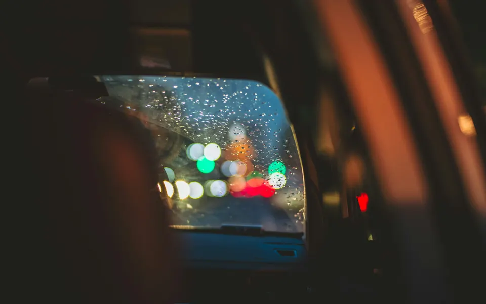 Blick aus dem dunklen Auto in den Seitenspiegel, Regentropfen und verschwommene Lichter der Straße