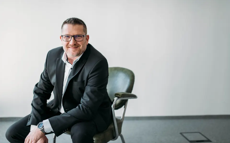 Andreas Pirker sitzt in hellem Raum auf einem Stuhl und lacht in die Kamera