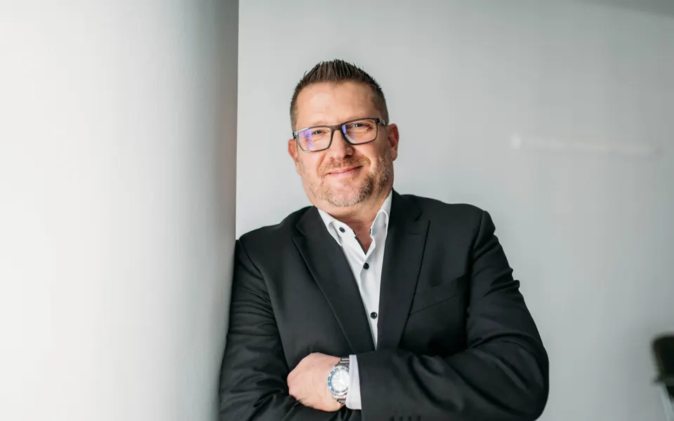 Andreas Pirker, Versicherungsmakler, lehnt mit verschr&auml;nkten Armen an einer hellen S&auml;ule und l&auml;chelt in die Kamera
